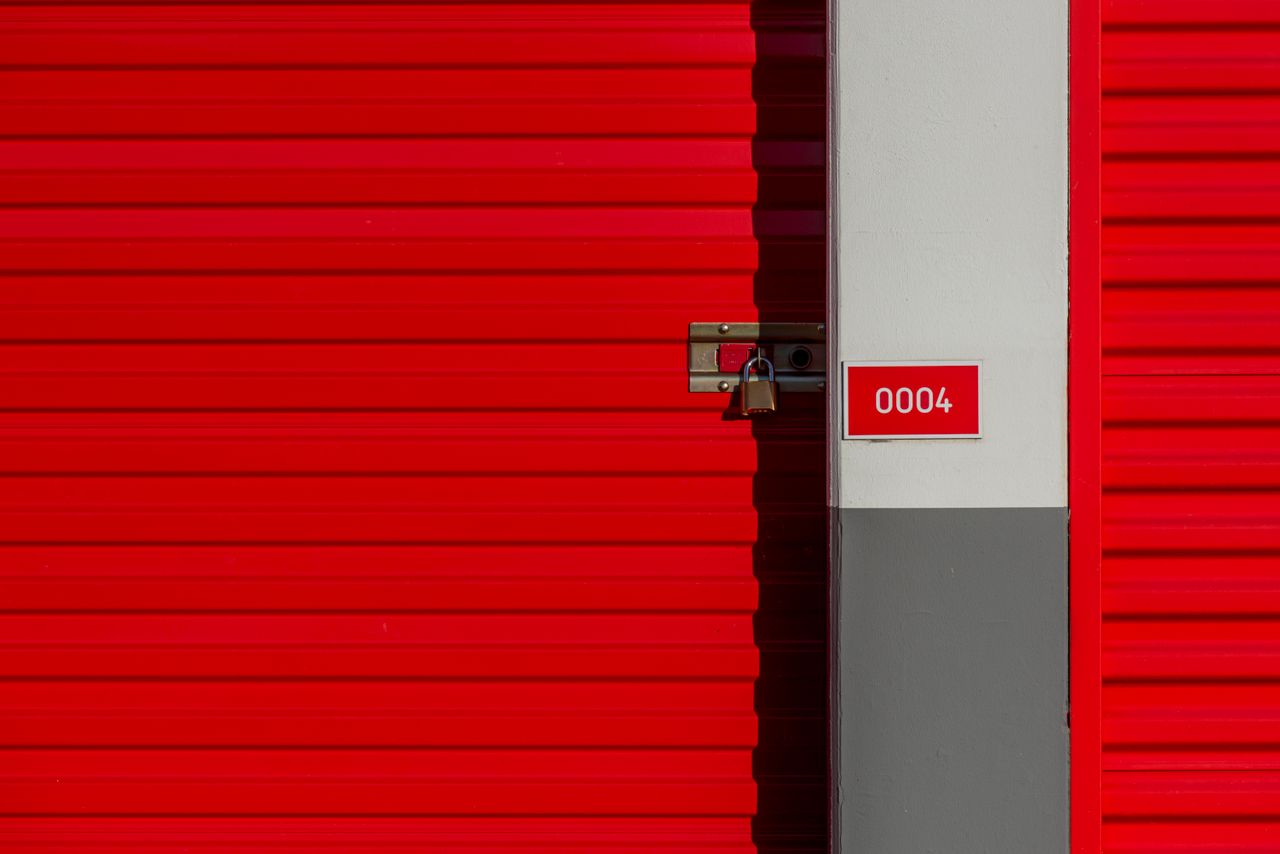 Red metal storage unit door with padlock and number 0004 sign on white panel.